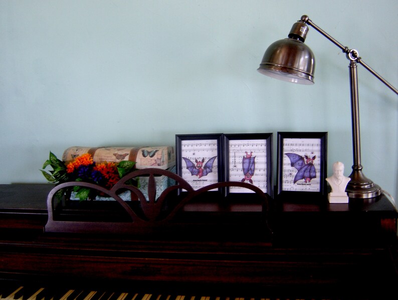 May include: Three framed prints of bats with musical notes in the background. The prints are on a wooden surface with a lamp and a decorative box with flowers.