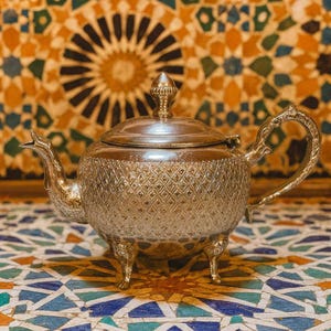 Peut inclure: Théière argentée avec un motif en losanges, une anse incurvée et un couvercle décoratif. La théière repose sur trois petits pieds. Le fond présente un motif de carreaux de mosaïque colorés.