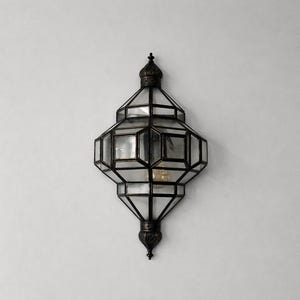 May include: An ornate, geometric wall sconce with a dark metal frame and clear glass panels. The fixture has a multi-faceted design, with a bulb visible inside. It is mounted on a white wall, showcasing its decorative style.