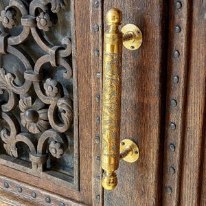 May include: A close-up of a brass door handle with an ornate floral design. The handle is mounted on a wooden door with visible grain and metal studs. The handle's finish is a bright gold color.