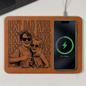 May include: A brown wireless charging pad with the text "BEST DAD EVER" and a photo of two children wearing sunglasses. A black smartphone is charging on the pad, displaying a charging symbol and 67% charged.