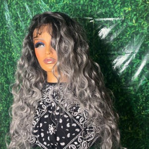 May include: A mannequin head displaying a long, wavy wig with a blend of gray and black tones. The wig is styled with voluminous curls. The mannequin is wearing a black and white bandana. The background is a green leafy pattern.