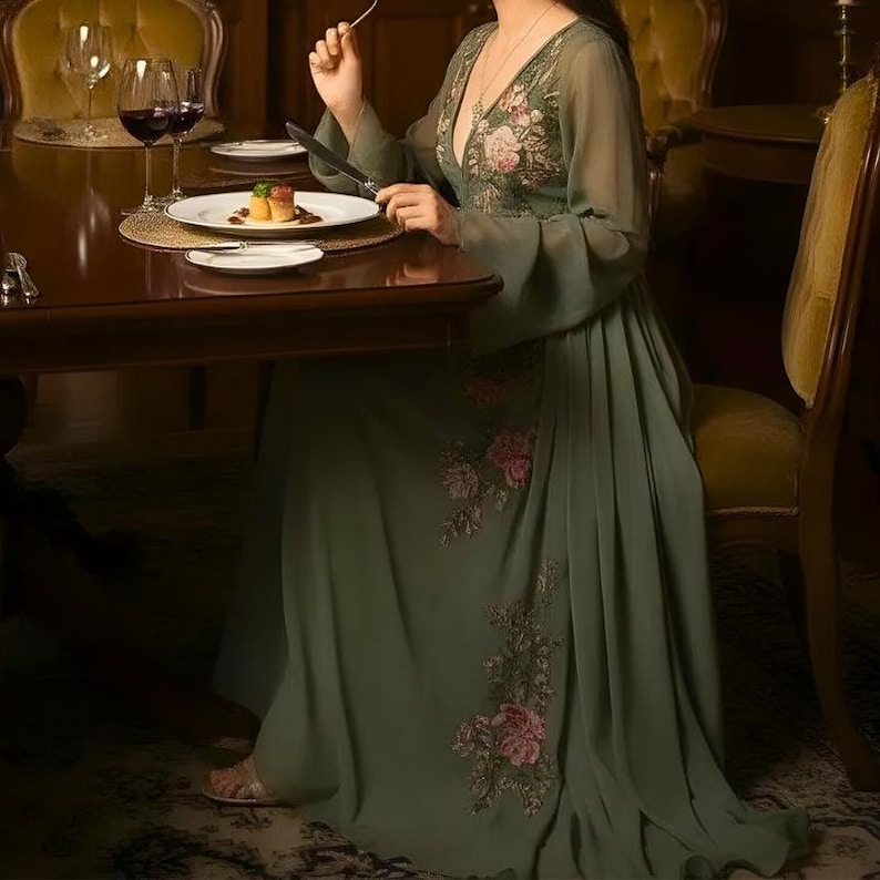 May include: An olive green formal gown with floral embroidery on the bodice and skirt. The dress features long, flowing sleeves and a deep V-neckline. The person is seated at a dark wooden table, with a plate of food and wine glasses visible.