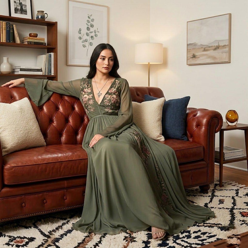 May include: An olive green maxi dress with floral embroidery on the bodice and sleeves. The dress features a V-neckline and flowing skirt. The model is sitting on a brown leather sofa. The dress is perfect for a special occasion.
