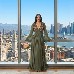 May include: A long, olive green dress with long, flowing sleeves and floral embroidery. The dress is worn by a person standing in front of a large window overlooking a cityscape. The dress has a V-neckline and a flowing skirt.