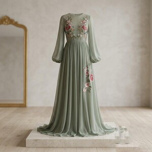 May include: A sage green floor-length dress with long, sheer sleeves and a flowing skirt. The dress features delicate floral embroidery in pink and white on the bodice and skirt. The dress is displayed on a white platform.