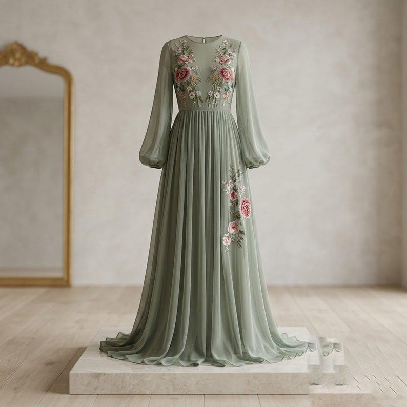 May include: A sage green floor-length dress with long, sheer sleeves and a flowing skirt. The dress features delicate floral embroidery in pink and white on the bodice and skirt. The dress is displayed on a white platform.