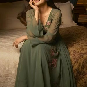 May include: A flowing sage green dress with long, sheer sleeves and a V-neckline. The bodice and sleeves feature delicate floral embroidery in shades of pink and red. The dress is made of a lightweight fabric, perfect for a special occasion.