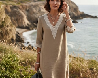 May include: A tan linen dress with a V-neckline trimmed with white lace. The dress has three-quarter sleeves with matching lace cuffs. The person is holding a gray hat and wearing layered necklaces and bracelets. The background features a coastal landscape.