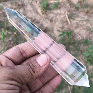Op de afbeelding: Een heldere, transparante, dubbelzijdige kristallen staaf met een gefacetteerde, zeshoekige vorm. Het object heeft gladde, gepolijste oppervlakken die licht reflecteren en loopt aan beide uiteinden uit in scherpe punten.
