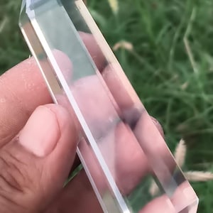 May include: A clear, transparent, double-terminated quartz crystal point with a smooth, polished, hexagonal faceted surface. The elongated, six-sided prism shape tapers to sharp, pointed ends on both sides, reflecting light through its body.