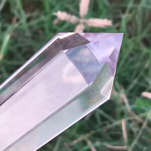 Op de afbeelding: Een heldere, zeszijdige, gefacetteerde kristallen punt met een scherpe top. Het kristal is transparant met een subtiele lichtpaarse tint en heeft gladde, gepolijste oppervlakken die het licht reflecteren langs de langwerpige, geometrische, hexagonale structuur.