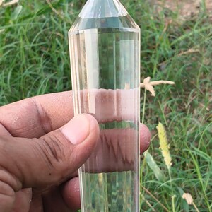 Può includere: Una punta di cristallo di quarzo trasparente a doppia terminazione con una superficie sfaccettata esagonale liscia e lucida. Il cristallo è allungato e presenta estremità appuntite sia nella parte superiore che inferiore.