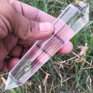 Può includere: Una bacchetta di cristallo di quarzo trasparente a doppia terminazione con una struttura esagonale sfaccettata. La pietra traslucida presenta superfici lisce e levigate che si restringono in punte affilate su entrambe le estremità.