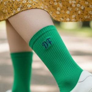 Può includere: Una persona indossa calzini verdi con le lettere "DF" ricamate in blu navy, abbinati a scarpe da ginnastica bianche. La persona indossa anche un vestito giallo a fiori. L'immagine è scattata all'aperto su un sentiero lastricato.
