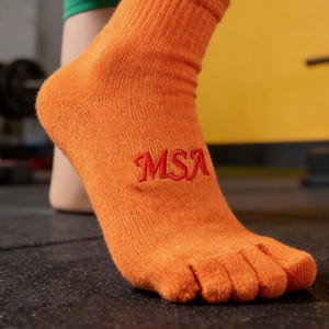May include: Orange toe socks with the embroidered letters "MSA" in red. The socks are made of a ribbed knit material and have individual compartments for each toe. The background is a blurred view of a gym setting.