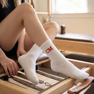 Op de afbeelding: Witte sportsokken met de rode initialen "CM" zijn zichtbaar. De sokken hebben een getextureerde zool en worden gedragen tijdens het gebruik van een Pilates reformer machine. De sokken zijn gemaakt van een zacht, comfortabel materiaal.