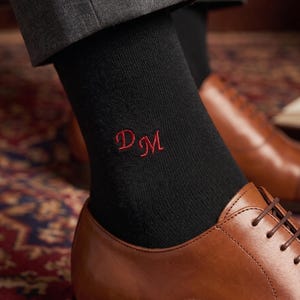 May include: Black socks with red embroidered initials "DM" are paired with brown leather dress shoes. The socks are visible above the shoes, and the person is wearing gray trousers. The shoes have a classic lace-up design.