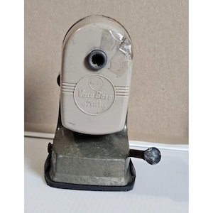 Op de afbeelding: Vintage Vacu Base puntenslijper in grijstinten. De puntenslijper heeft een handslinger en de tekst "Vacu Base Pencil Sharpener" is zichtbaar. Gemaakt van metaal en plastic. Een klassiek kantoorartikel.