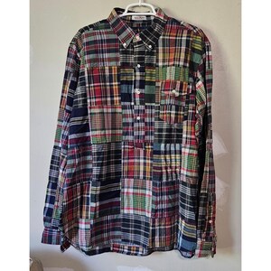 May include: A long-sleeve button-down shirt featuring a multi-colored patchwork plaid pattern. The shirt has a buttoned collar, a single chest pocket on the left side, and a partial button placket extending from the collar to the mid-chest area.