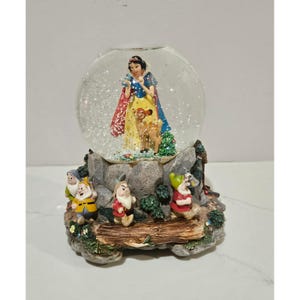 May include: A Snow White snow globe featuring a glass sphere containing a Snow White figurine and a deer. The base is a rocky landscape with three dwarf figurines, green foliage, and a wooden log detail. The globe is filled with white glitter particles.