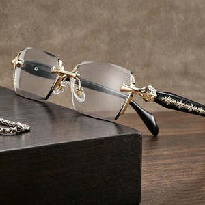 Könnte beinhalten: Rahmenlose Sonnenbrille mit goldfarbenen Akzenten und schwarzen Bügeln. Die Gläser sind leicht grau getönt. Die Bügel weisen ein dekoratives Muster auf. Die Sonnenbrille liegt auf einer dunklen Holzoberfläche.