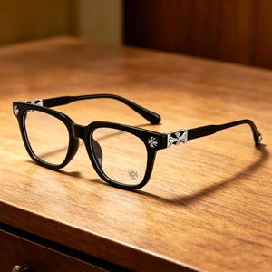 Könnte beinhalten: Schwarze Brille mit quadratischem Rahmen und klaren Gläsern. Die Bügel und Rahmenecken weisen silberne Kreuz- und Blumenmotive auf. Die Brille liegt auf einer Holzoberfläche.