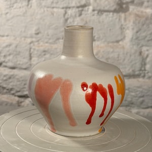Könnte beinhalten: Eine Keramikvase mit einer glatten, cremeweißen Basis, die vertikale, tropfenförmige Glasurmuster in den Farben zartes Rosa, leuchtendes Rot und helles Orange aufweist. Das Gefäß hat einen abgerundeten Körper und einen schmalen Hals.