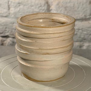 Könnte beinhalten: Ein zylindrischer Keramikbecher mit einer hellbeigen, cremeweißen Glasur. Die Außenfläche weist eine Reihe von horizontalen, erhabenen Rillen oder Wülsten auf, die sich um den Körper ziehen und ein strukturiertes, geschichtetes Erscheinungsbild erzeugen.