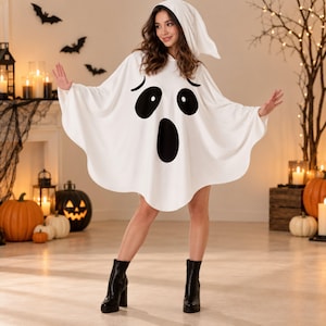 May include: A white hooded poncho featuring a large black ghost face print with two oval eyes and an elongated oval mouth. The garment has a loose, draped silhouette with wide sleeves and a pointed hood, creating a classic ghost costume appearance.