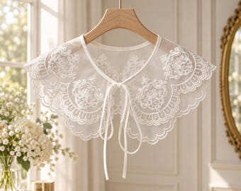 May include: A white detachable collar made of sheer fabric featuring intricate floral embroidery and scalloped edges. The collar has a rounded shape and includes two long, thin white fabric ties at the center front for closure, hanging from a wooden hanger.