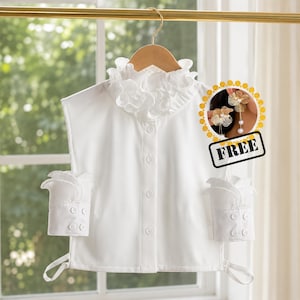 May include: A white, sleeveless blouse with a ruffled collar and cuffs, displayed on a wooden hanger. The cuffs have button details. A small image of earrings with the word "FREE" is in the corner.