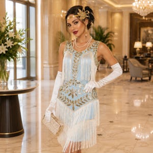 May include: A light blue sleeveless dress featuring gold sequin leaf patterns and long white fringe layers. The ensemble includes white elbow-length satin gloves, a multi-strand pearl necklace, and a gold decorative headpiece with a floral design.