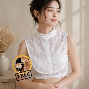 May include: White sleeveless blouse with a high, ruffled collar and floral cut-out details. The blouse has a button-down front and a lace trim at the bottom. The image also includes a pair of gold-toned earrings with pearl accents and the word "FREE".