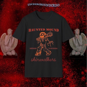 May include: Black t-shirt with orange graphic and text. The graphic features a figure with a pumpkin head holding a weapon. Text includes "HAUNTED MOUND", "HAUNTED MOUND", "TILL IN THE GROUND", and "skinwalkers". The shirt is displayed with angel statues.