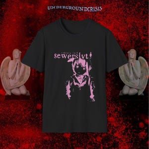 May include: Black t-shirt with a pink graphic of a stylized anime character and the text "sewerslvt" above the image. The text "UNDERGROUNDCRISIS" is at the top. Two angel statues flank the shirt.