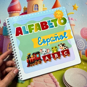 Op de afbeelding: Een vierkant notitieboek met spiraalbinding en een lichtblauwe kaft met de tekst ALFABETO Español in kleurrijke letters. De kaft bevat een gele zon, een rode treinlocomotief en drie treinwagons met een grijze olifant, een groene kikker en een koe.