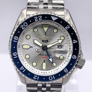 Może przedstawiać: Zegarek automatyczny Seiko 5 GMT ze stali nierdzewnej ze srebrną tarczą, świecącymi znacznikami i niebiesko-czarnym pierścieniem. Zegarek posiada wyświetlacz daty i bransoletę ze stali nierdzewnej.