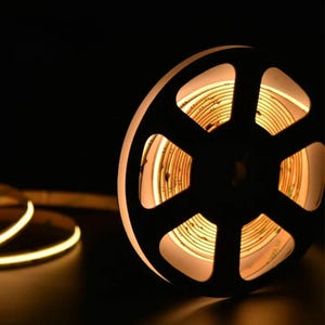 May include: A roll of warm white LED strip lights, illuminated against a dark background. The spool is black with a circular design, and the light strip is coiled inside. A section of the strip extends out, showcasing the flexible design.