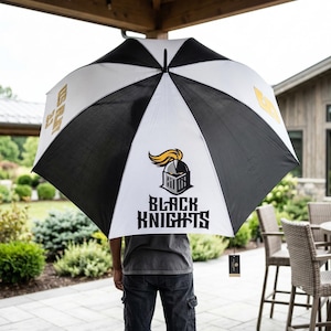 May include: A large, open umbrella with alternating black and white panels. The umbrella features the text "BLACK KNIGHTS" and a logo with a knight's helmet and a gold plume. The umbrella is held open.