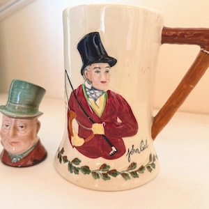 May include: A cream-colored ceramic mug and a small figurine. The mug features a man in a red coat and black top hat, with the name "John Peel" written below. The figurine is a man in a green top hat and red coat.