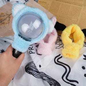 May include: A light blue plush light stick with white accents, a clear globe, and a black handle. Beside it are pink and yellow plush accessories. The light stick is held by a hand, and the background includes printed paper and a patterned fabric.