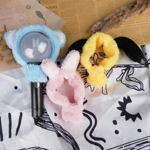 May include: Three plush light stick covers in the image. One is light blue with bear ears, one is pink with bunny ears, and the other is yellow with black wings. The light sticks are not included.
