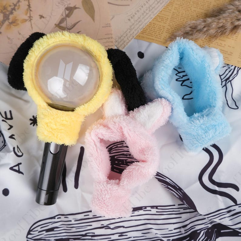 May include: Three plush light stick covers in yellow, pink, and blue. The yellow cover has black ear accents and a clear light. The pink cover has bunny ears. The blue cover has white ear accents. The light sticks are on a patterned surface.
