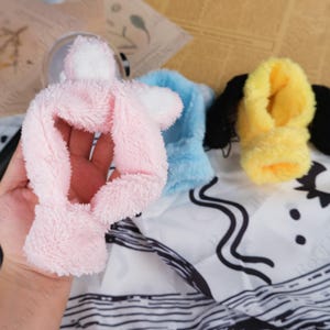 May include: A hand holds a soft, pink plush bunny ear headband with white accents. A light blue and yellow plush headband are also visible. The headbands are made of a fuzzy material.