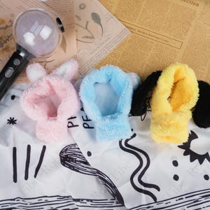 May include: Three plush light covers in pink, blue, and yellow, each with animal ear details. The covers are designed to fit over a light stick, with a black handle visible on the left. The items are on a white patterned surface.