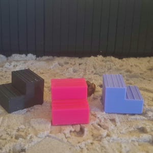 Puede incluir: Tres pequeños objetos en forma de escalón, en negro, rosa y azul, están dispuestos sobre una superficie arenosa. El objeto negro está a la izquierda, el rosa en el centro y el azul a la derecha. El fondo es un panel oscuro y texturizado.