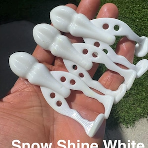 May include: Three white, glossy, semi-translucent plastic fishing lures with a segmented, elongated body shape. Each lure features a rounded head section, a slender midsection, and a flat, flared tail end with two circular cutouts in the body.