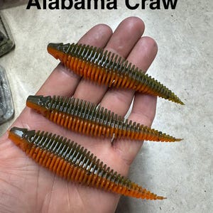 May include: Three soft plastic fishing lures labeled Alabama Craw. Each lure features a segmented, ribbed body with a dark olive green top and a bright orange bottom, tapering to a thin, pointed tail. The lures have a textured, worm-like design.