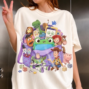 May include: Cream-colored t-shirt featuring a colorful graphic of characters from the animated movie Toy Story. The design includes Woody, Buzz Lightyear, and other characters inside a purple backpack. The shirt is a relaxed fit.